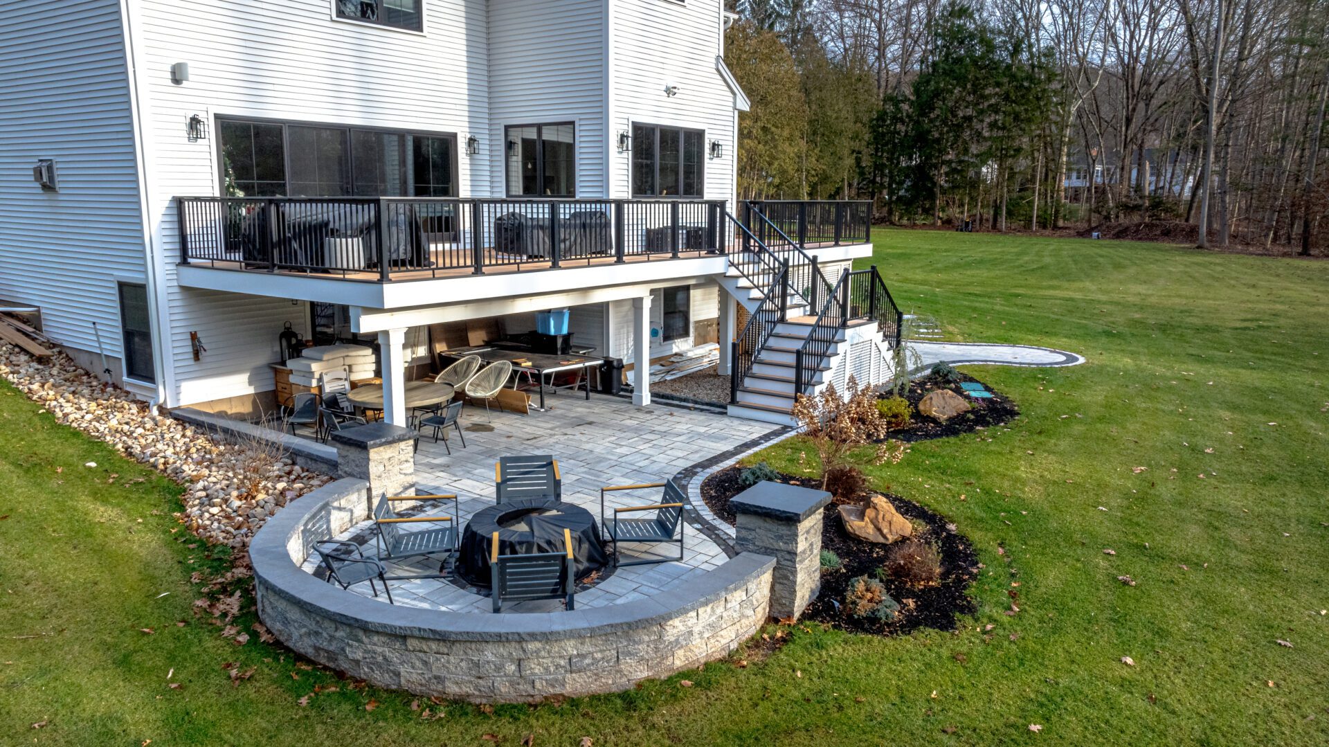 Side angle view highlighting an inviting stone patio with a semi-circular stone seating wall, integrated fire pit, and stylish landscaping beneath an elevated deck attached to a modern home.